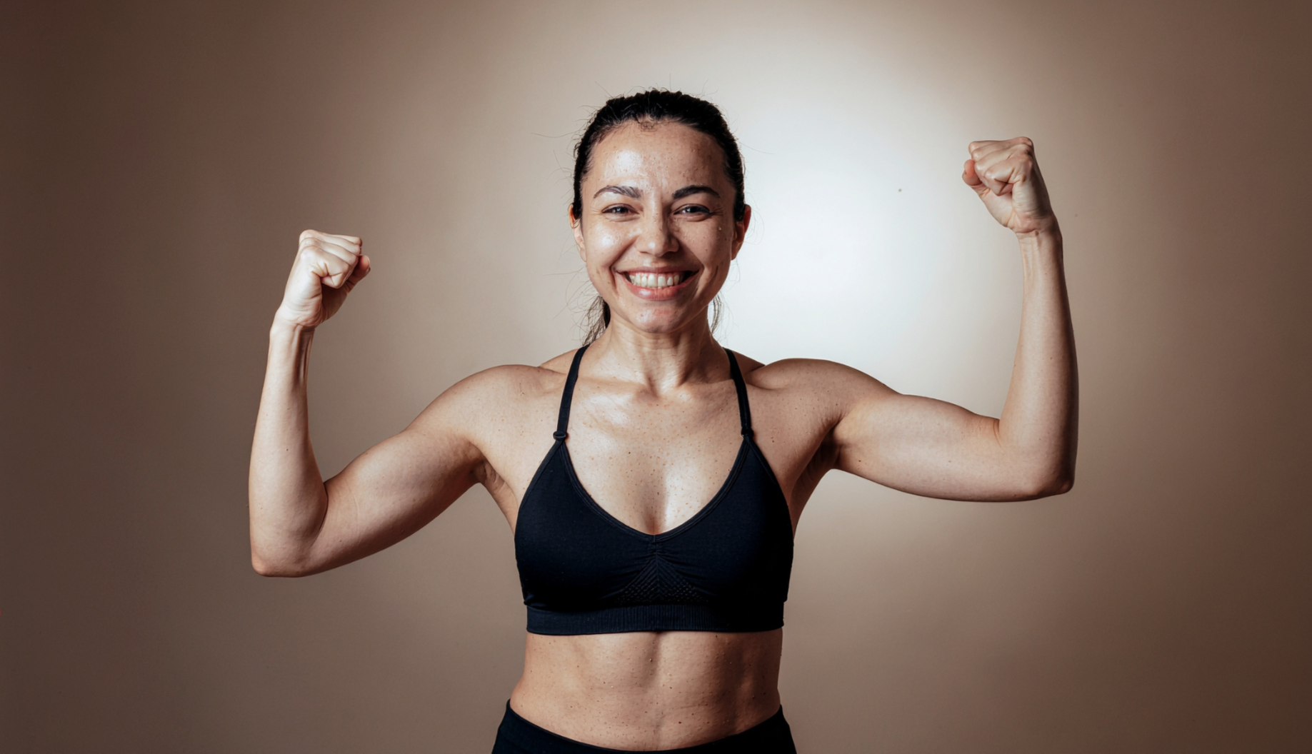 A woman flexing her biceps in a black sports bra, smiling confidently, with a plain beige background.