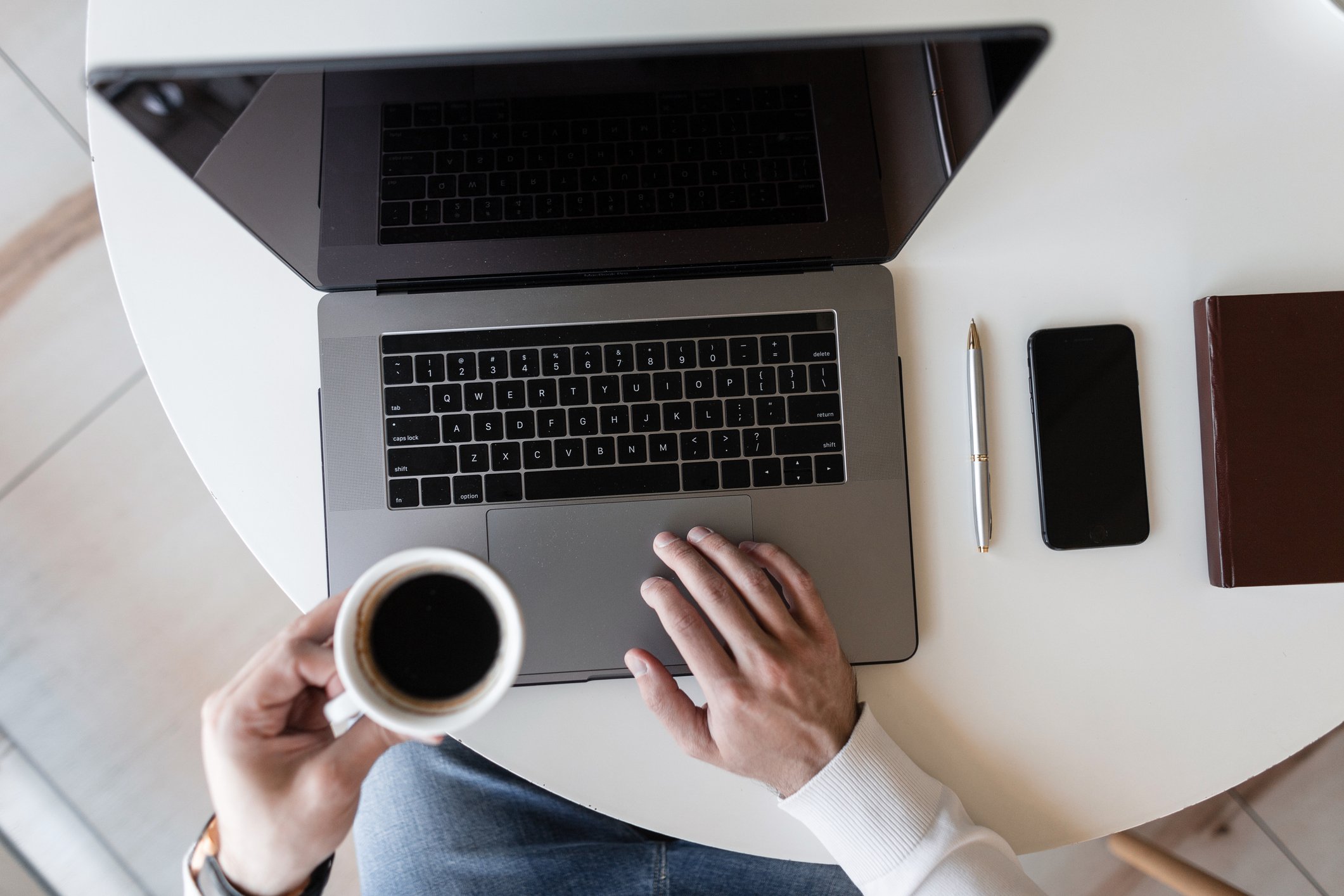 Top-down view of a person working on a laptop at a white table, holding a cup of coffee in their left hand, with a smartphone, pen, and closed notebook nearby.