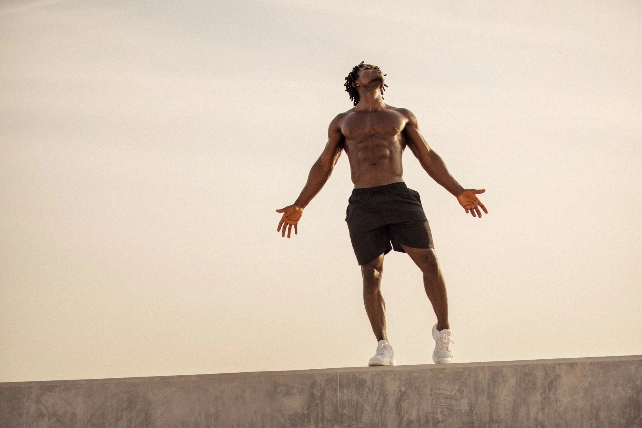 A shirtless man with dreadlocks standing outdoors on a ledge with arms outstretched, looking up with a serene expression.