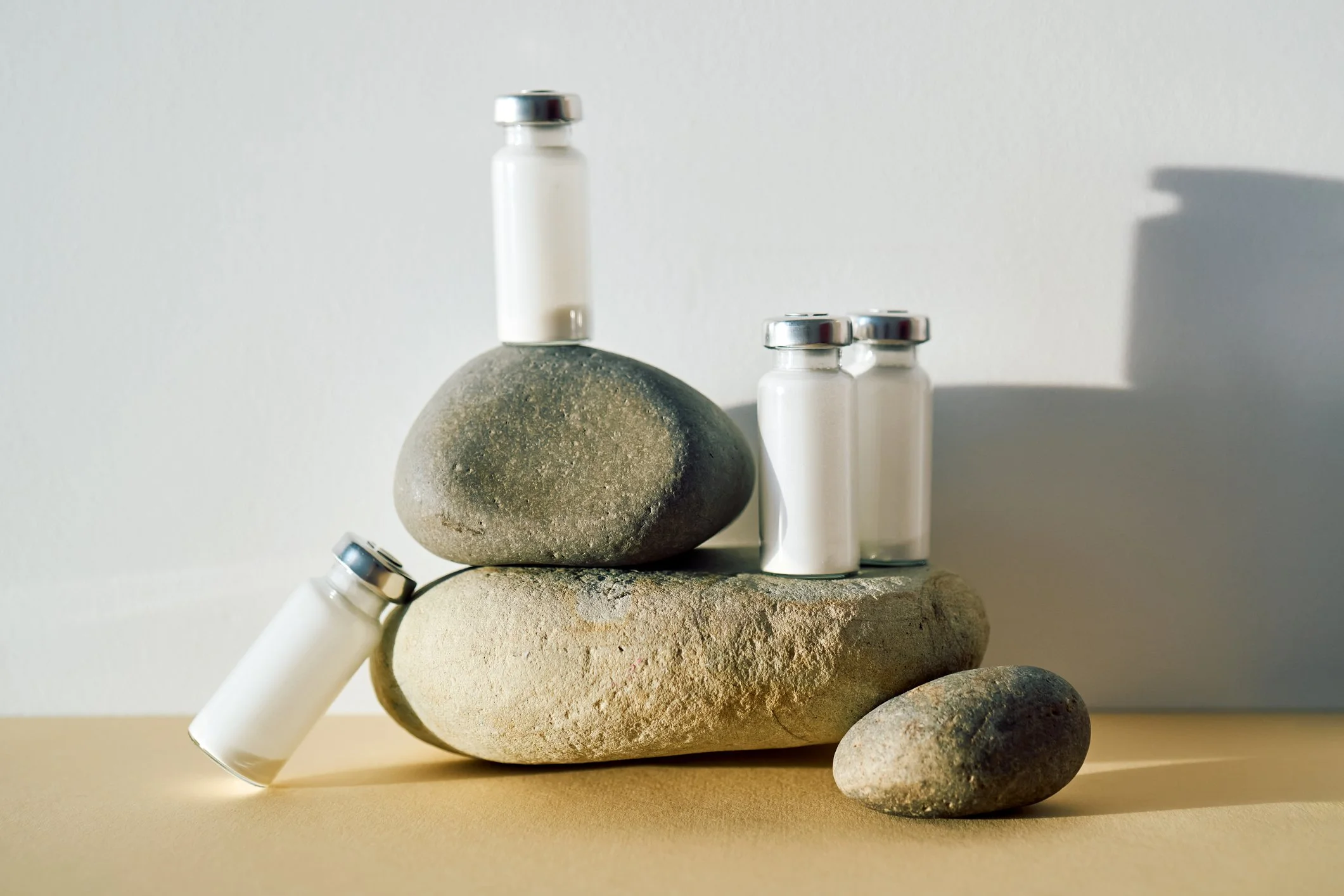 Multiple stones stacked with three small white bottles with silver caps placed on top and beside the stones, casting shadows on a plain light-colored background.