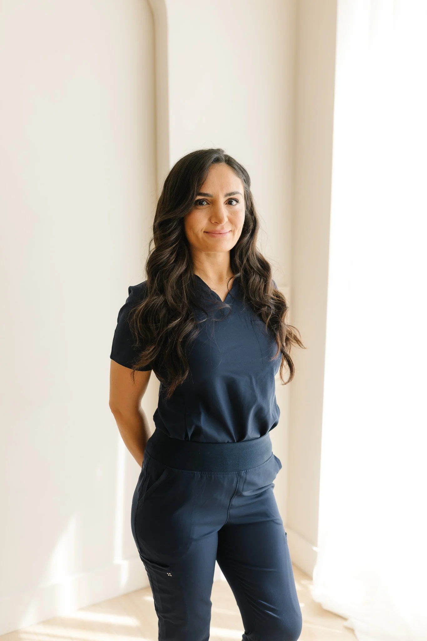 A woman with long dark hair wearing a navy blue scrubs top and matching navy pants stands in a well-lit room with white walls and a window with sheer curtains, smiling slightly at the camera.