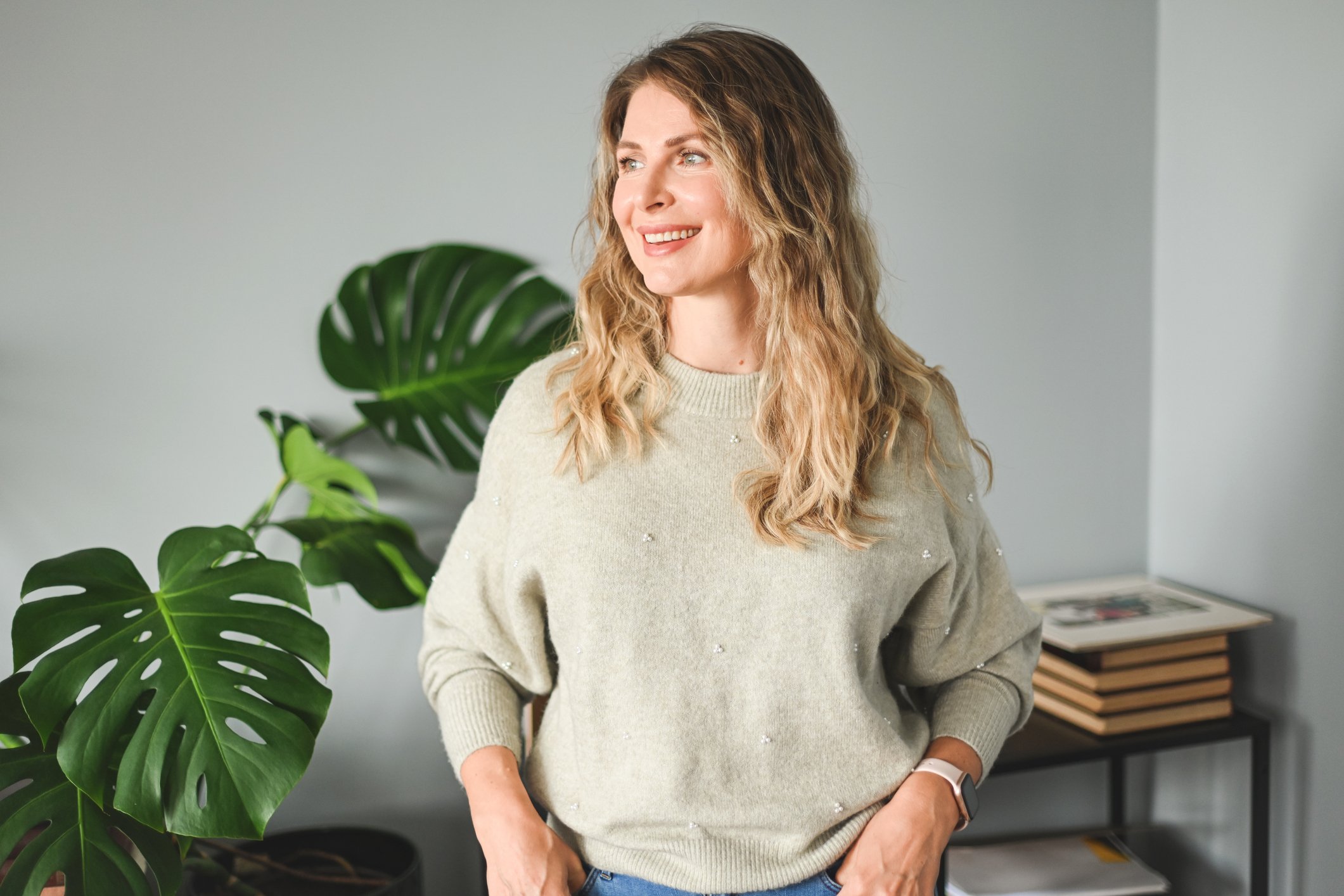A woman with wavy blonde hair smiling and looking to her right, standing inside a room with a large green plant nearby and a desk with magazines in the background.