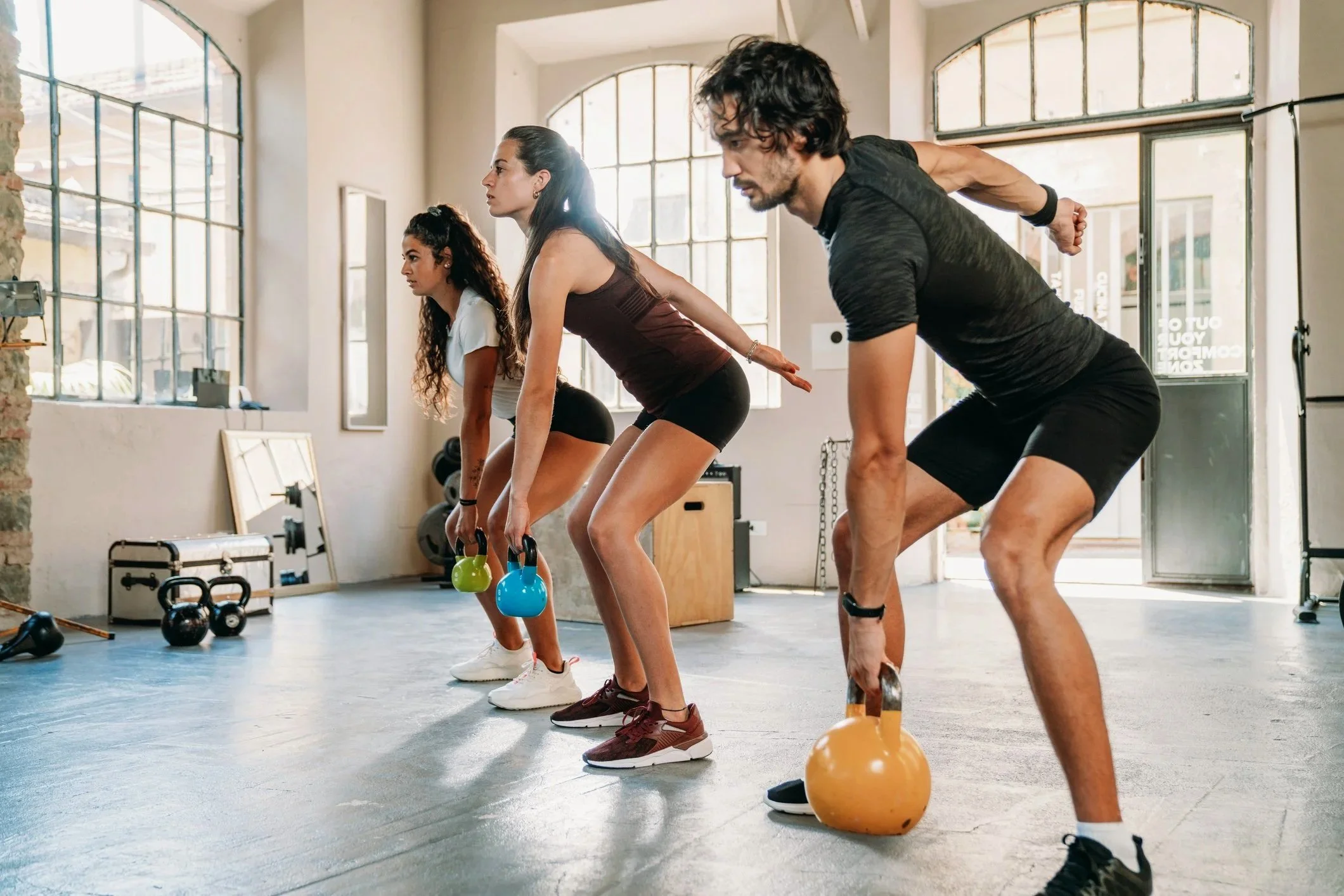 Four people in a gym doing a kettlebell workout together, facing downward, wearing athletic clothes, with gym equipment and large windows behind them.