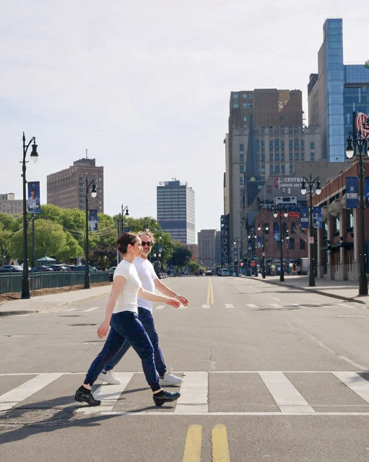 Two women walking across a city street on a sunny day, with tall buildings and trees in the background.