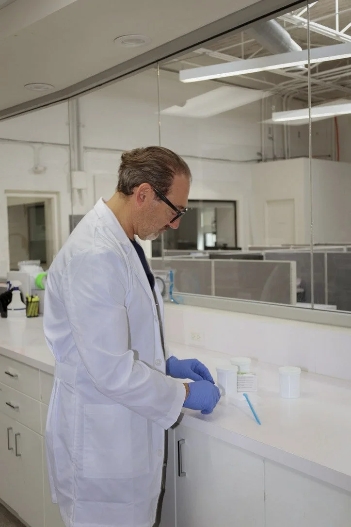 A scientist in a white lab coat and blue gloves preparing or examining a sample in a modern laboratory with glass partitioning and white cabinets.
