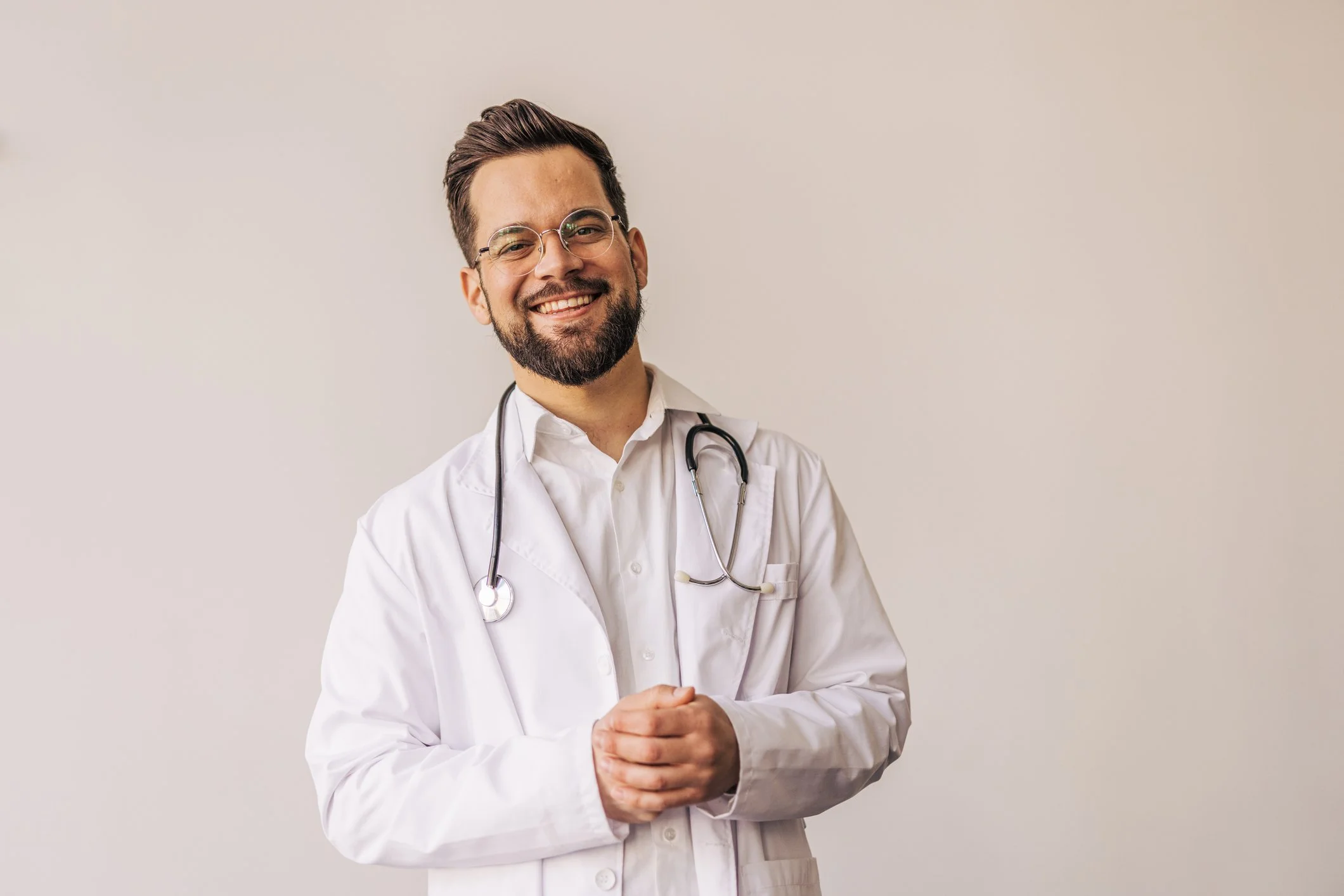 A smiling male doctor with glasses and a stethoscope around his neck, wearing a white coat, standing against a plain light-colored background.