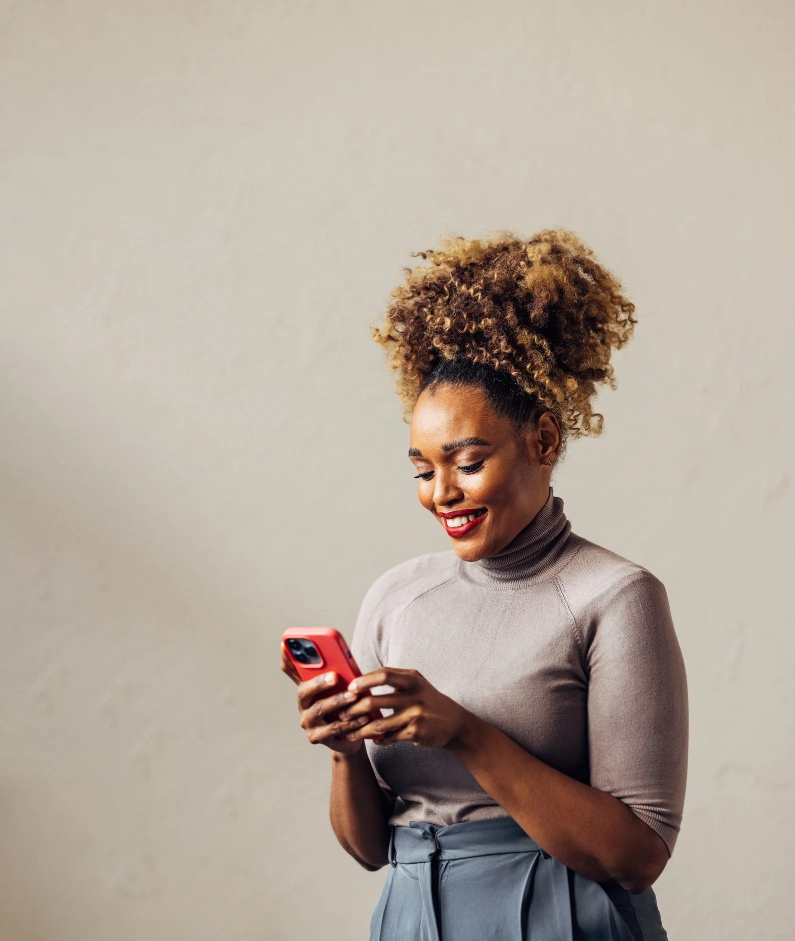 A woman with curly hair styled in a high puff, wearing a beige turtleneck sweater and gray skirt, smiling while looking at her phone against a plain beige wall.