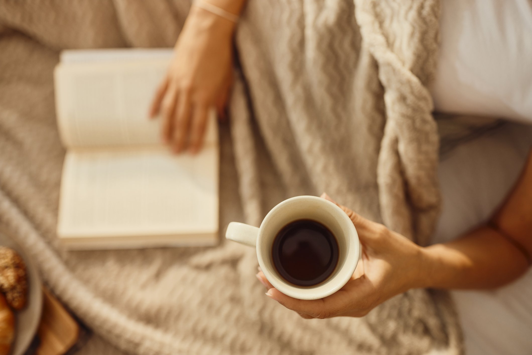 Top view of a person holding a white cup of black coffee over a kitchen countertop with a notebook, chain with a pendant, and some snacks.