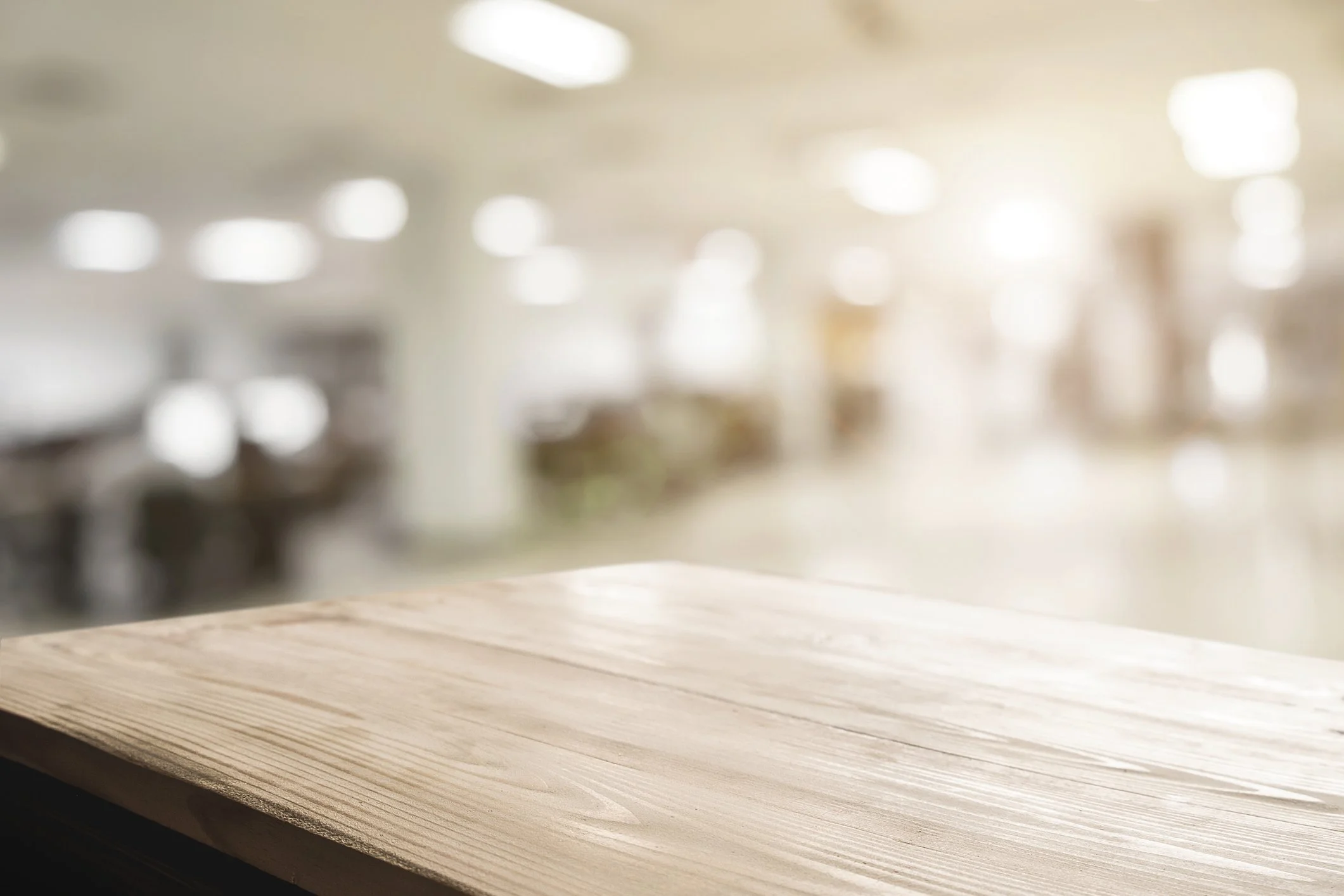A light wooden tabletop in focus with blurred background of a room, possibly a store or showroom, with soft overhead lighting.