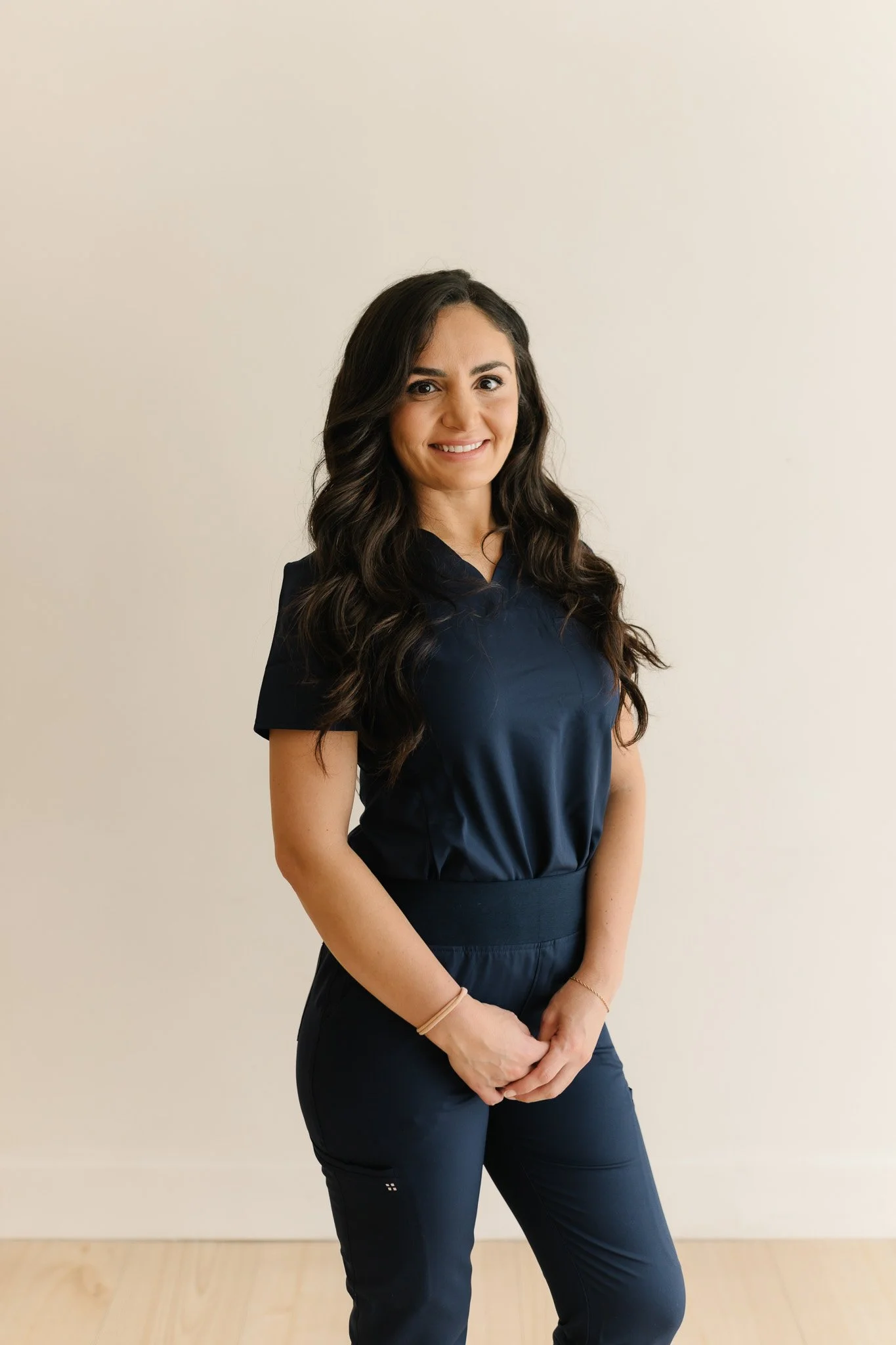 A woman with long dark wavy hair, smiling, dressed in a navy blue top and pants, standing against a plain beige wall.