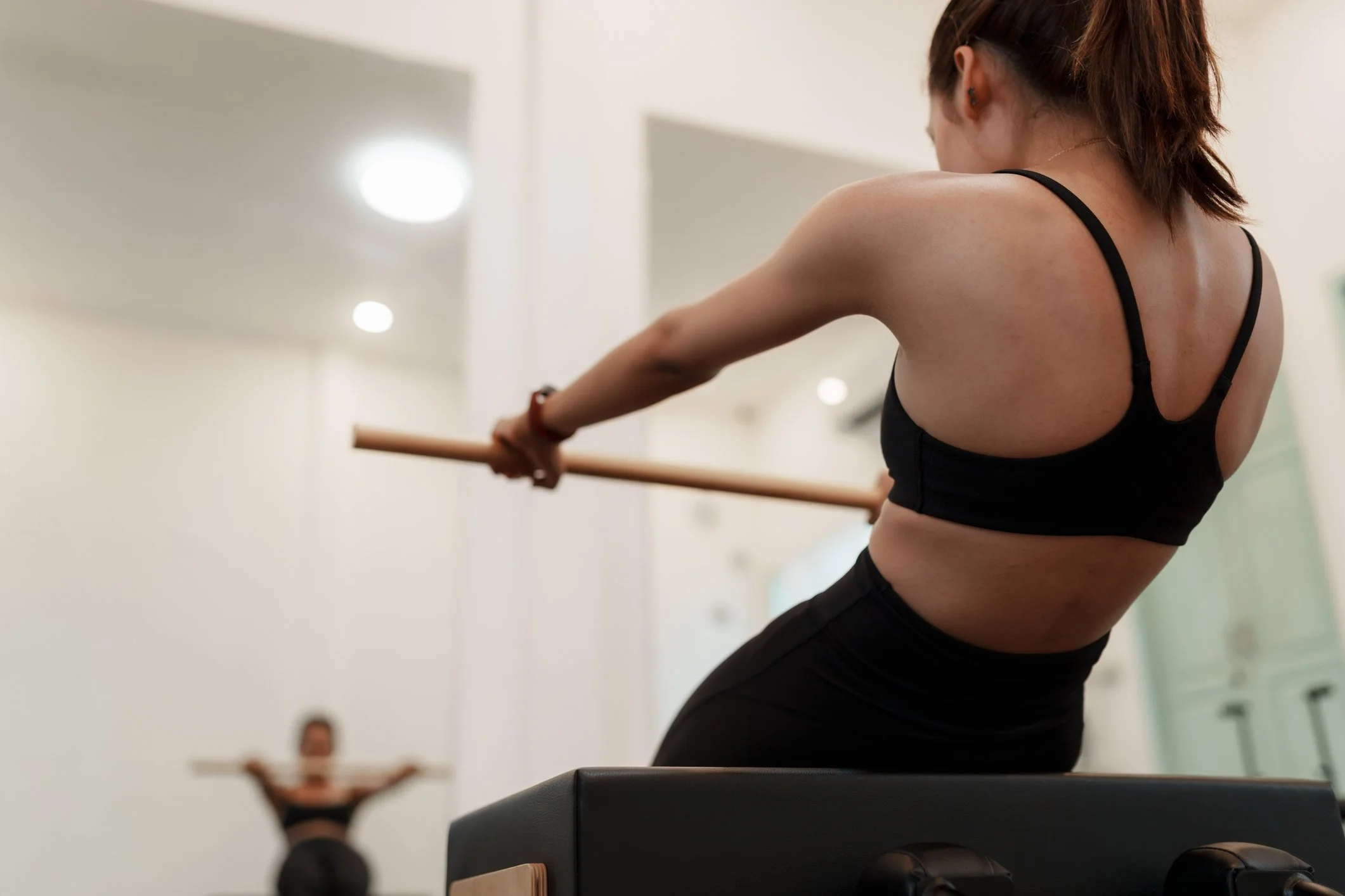 A woman in a black sports bra and black pants practicing ballet at a barre in a dance studio, with another woman in the background stretching or warming up.