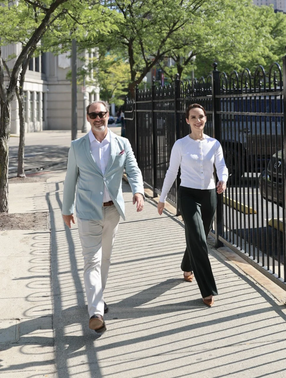 A man and woman walking on a sunny city sidewalk, smiling. The man is wearing sunglasses, a light blue blazer, white shirt, beige pants, and brown shoes. The woman is dressed in a white cardigan, black pants, and brown heels, with short dark hair. They are walking past a black metal fence with cars parked behind it, and green trees lining the street.