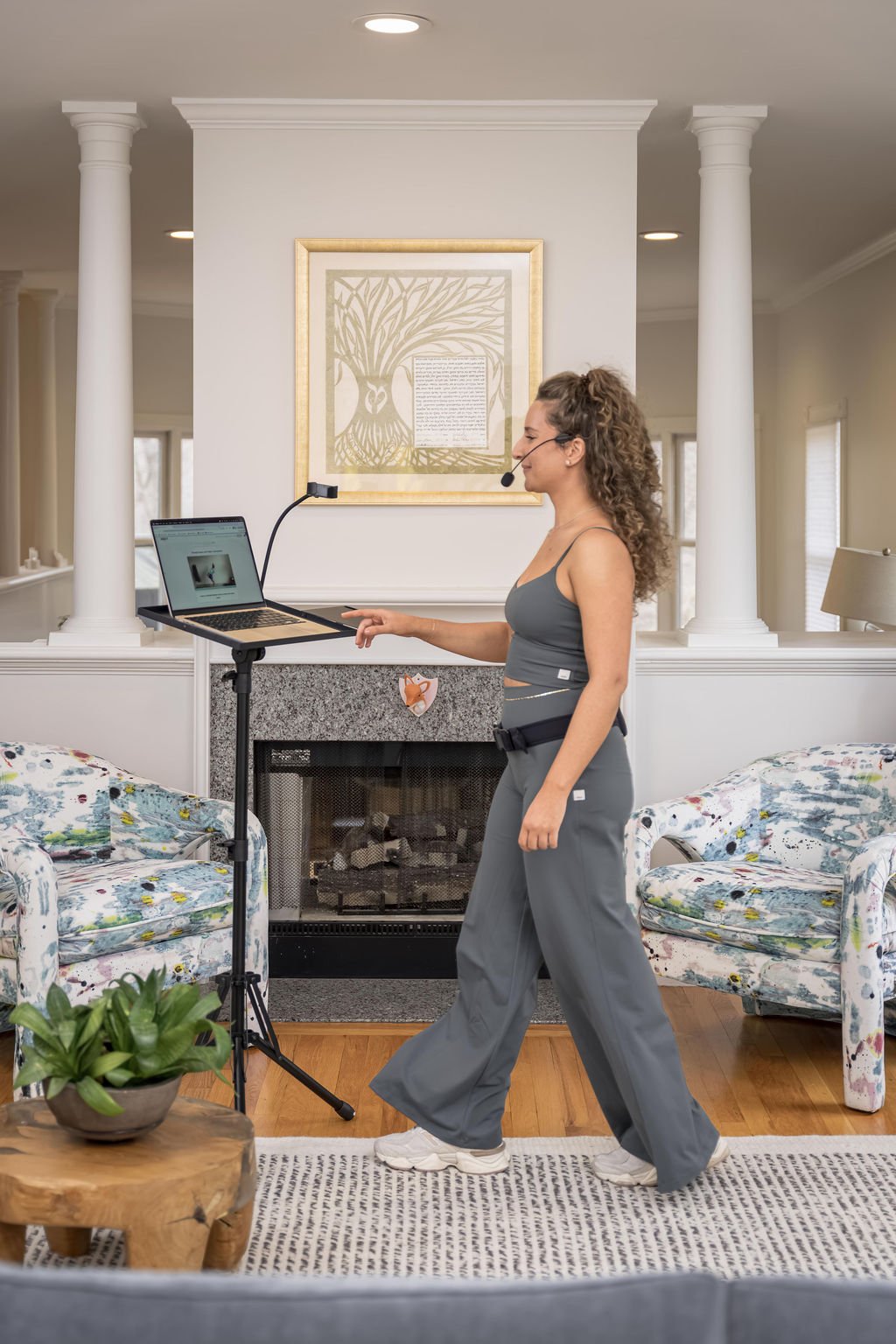 A woman with curly hair wearing a gray tank top and gray pants standing in a living room, using a standing desk with a laptop and a microphone headset.