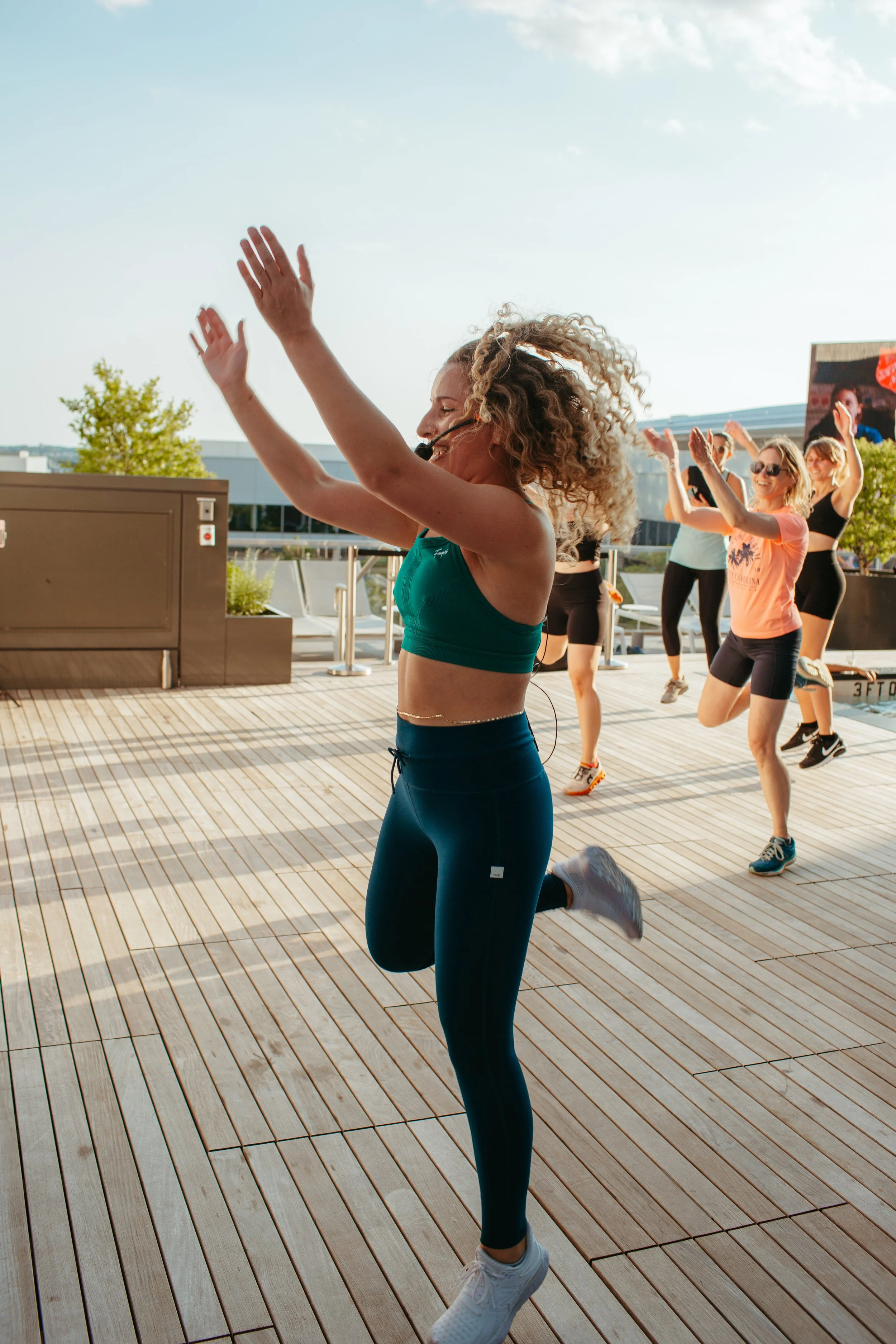Young women participating in an outdoor fitness class on a rooftop, with one instructor leading the exercise and others following, under a clear blue sky.