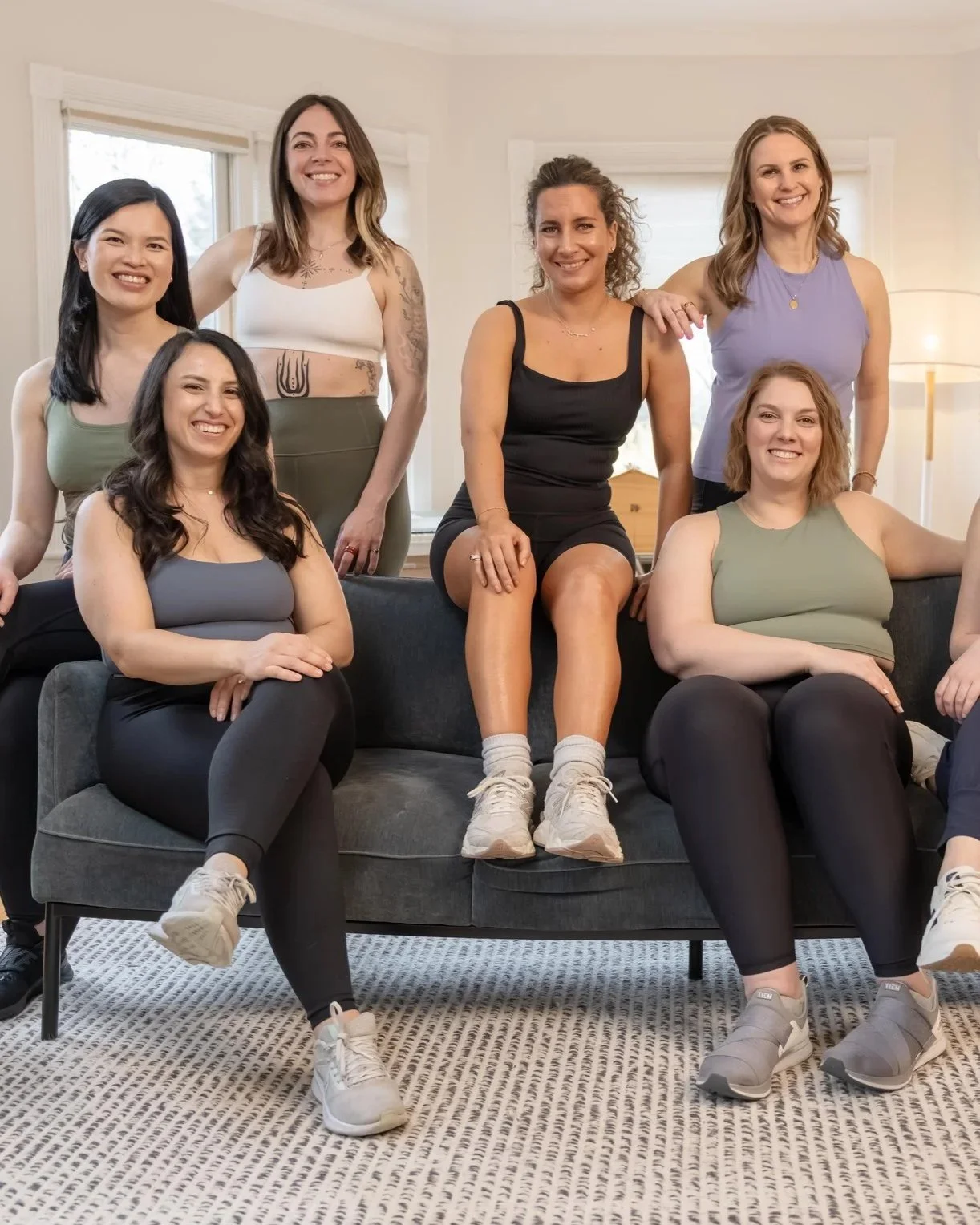 Group of six women sitting and standing on a dark grey sofa in a well-lit living room, smiling at the camera.