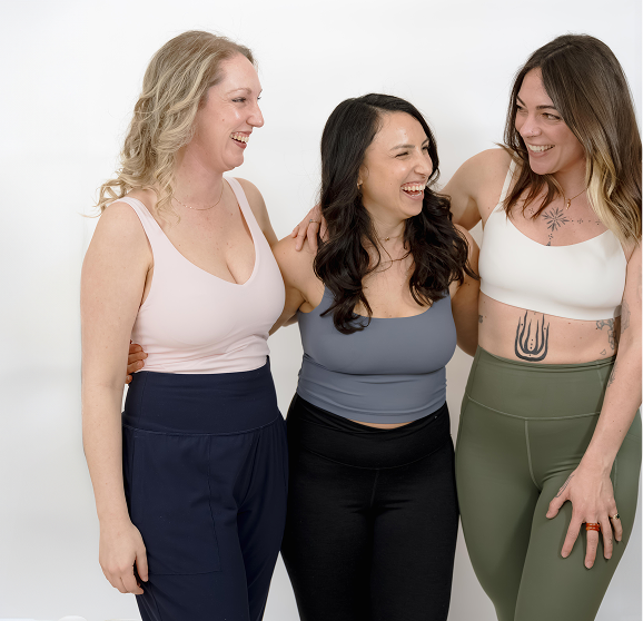 Three women smiling and laughing together in casual clothing against a white background.