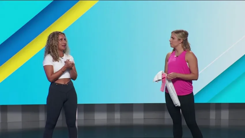 Two women talking on a colorful stage background, one in a white crop top and dark pants, the other in a pink sleeveless top and black pants, holding a towel.