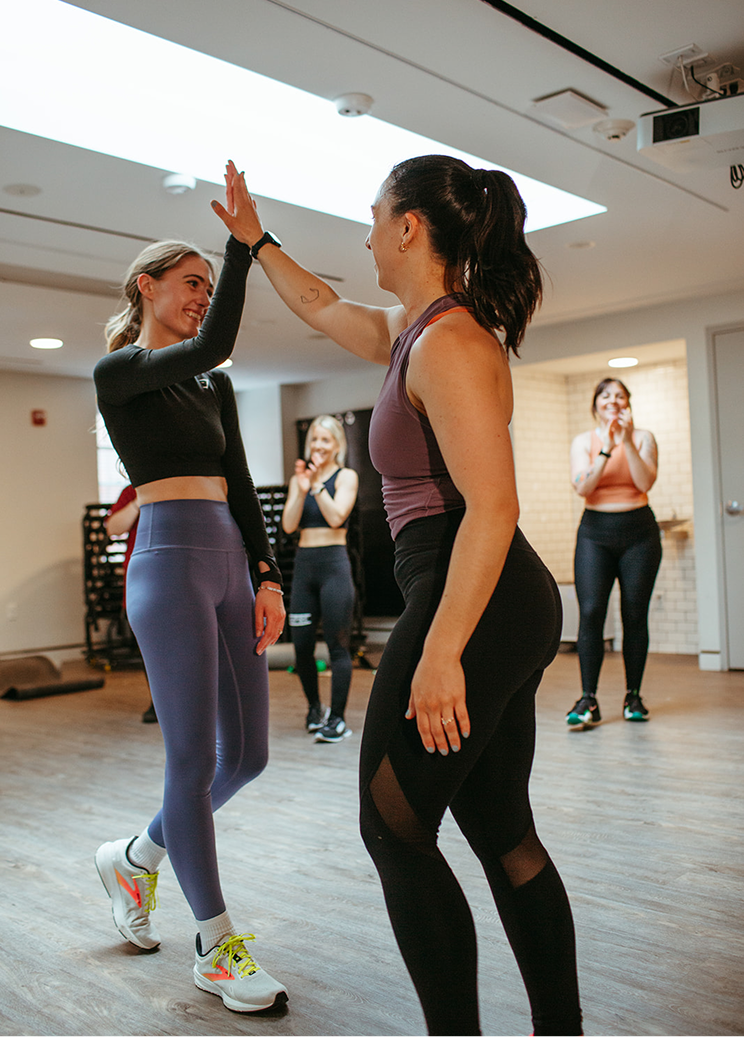 Two women high-five during a workout class, others clapping in the background, in a brightly lit gym.