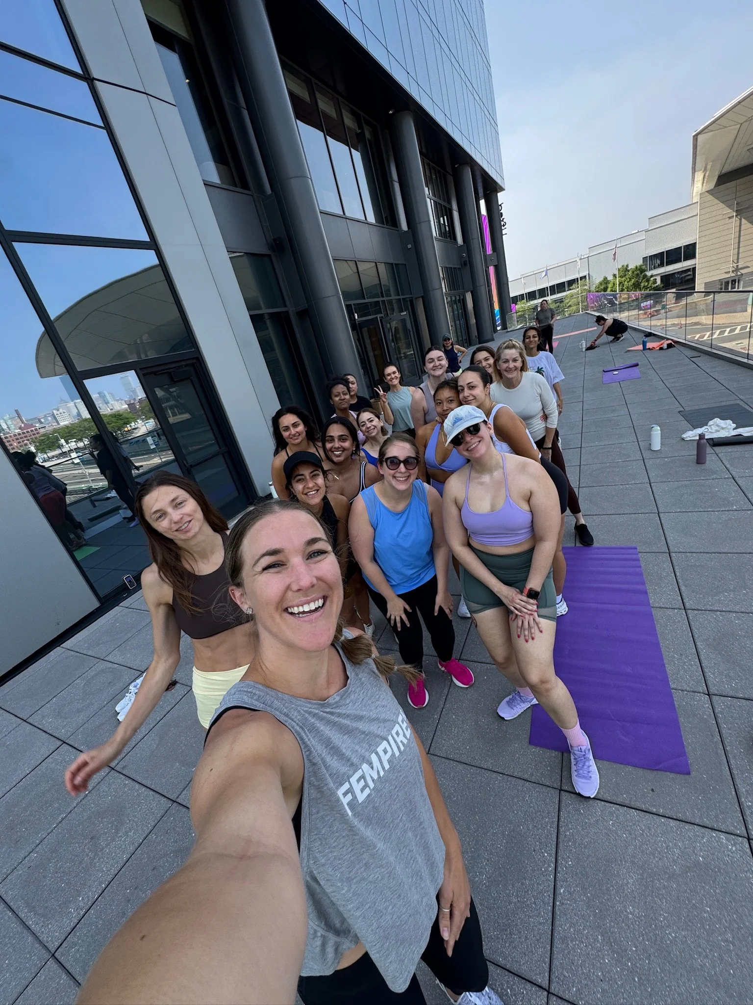 Group of women in workout clothes smiling and taking a selfie outside a modern building on a sunny day.