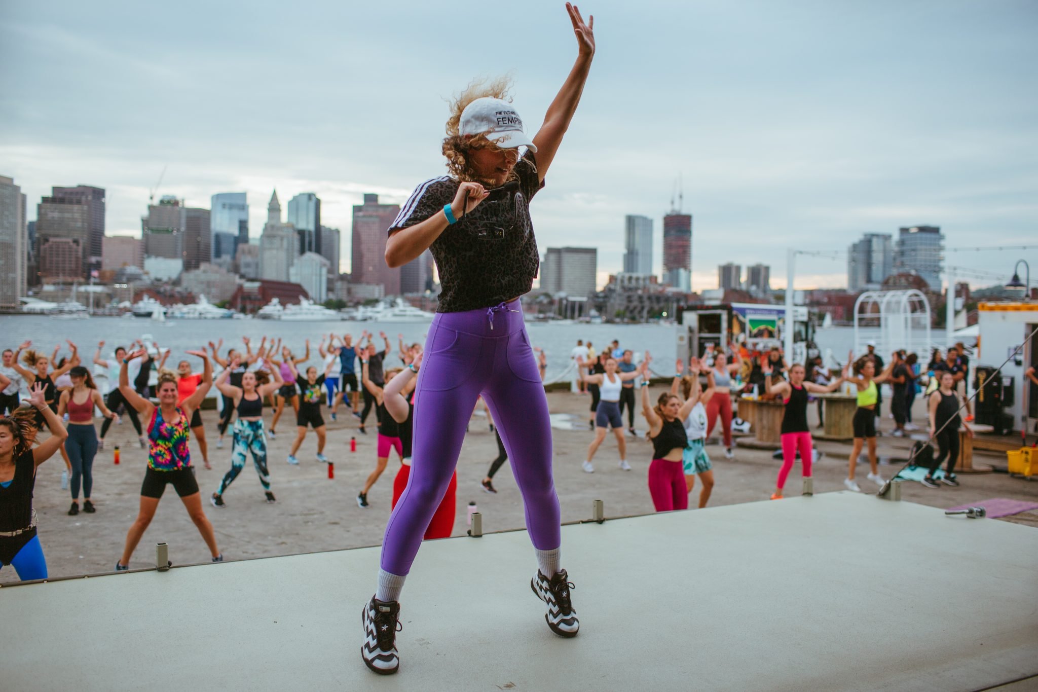 A woman leading an outdoor fitness class with many participants in a city waterfront area, practicing aerobics or dance exercises.