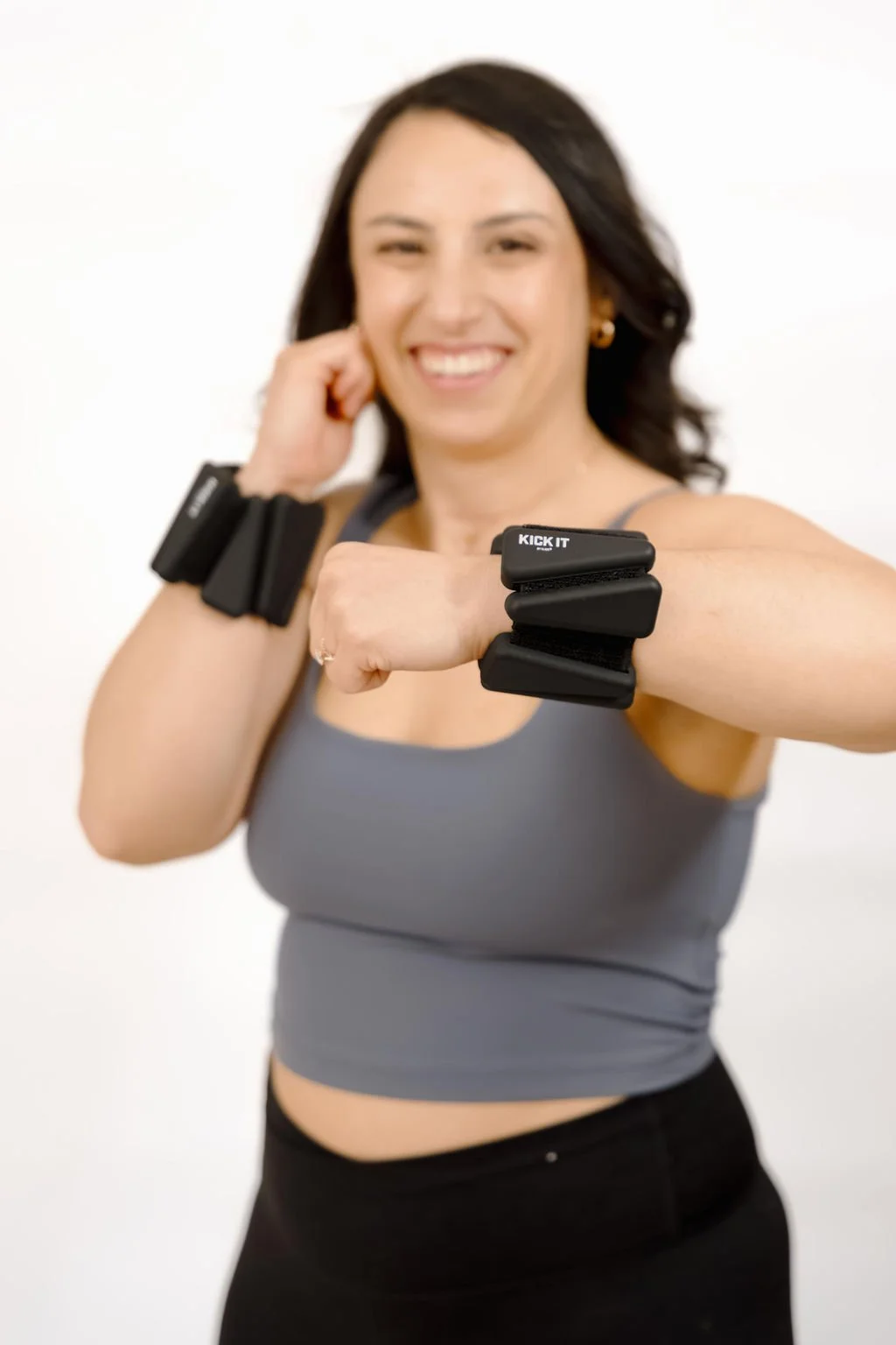 A woman smiling and wearing workout gear, showing off fitness equipment on her arm, including black wrist weights and a fitness tracker.