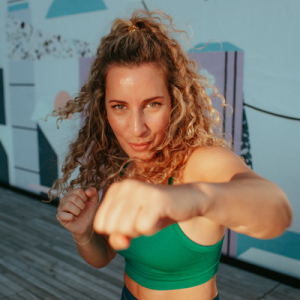 A woman with curly hair in workout attire punching towards the camera during a workout session outdoors.
