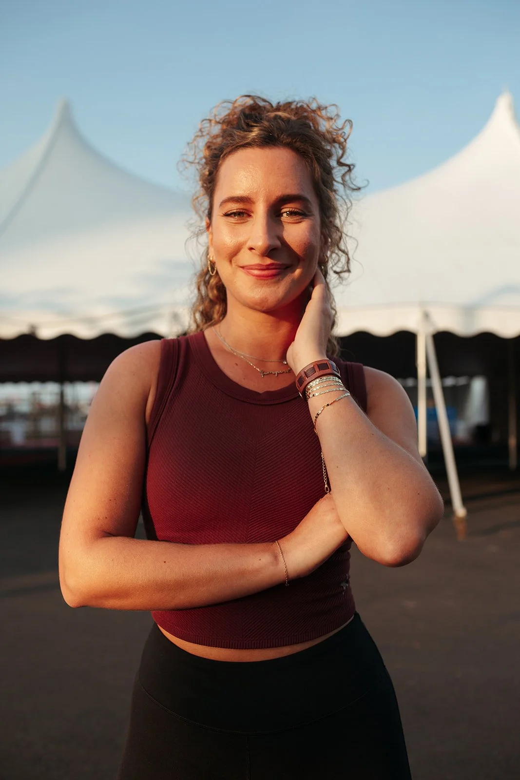 A woman smiling outdoors in front of white tents during sunset, wearing a sleeveless maroon top and jewelry.