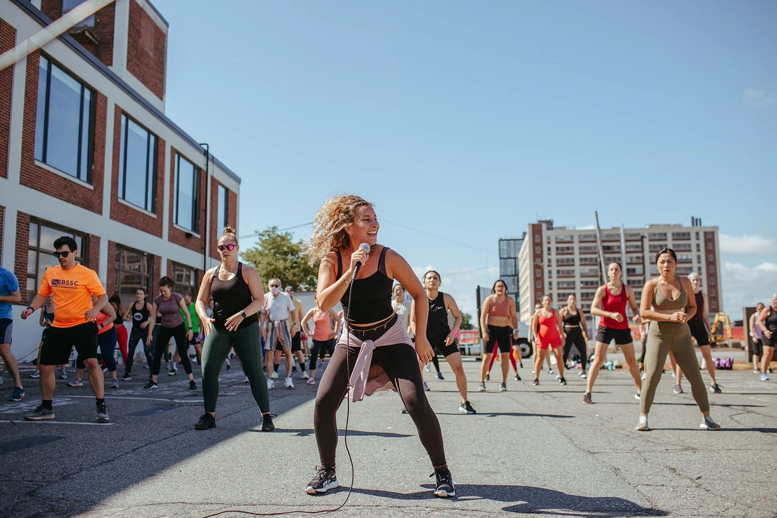 A woman leading a dance or fitness class outdoors with a microphone, surrounded by a group of people participating in the class on a sunny day in an urban area.