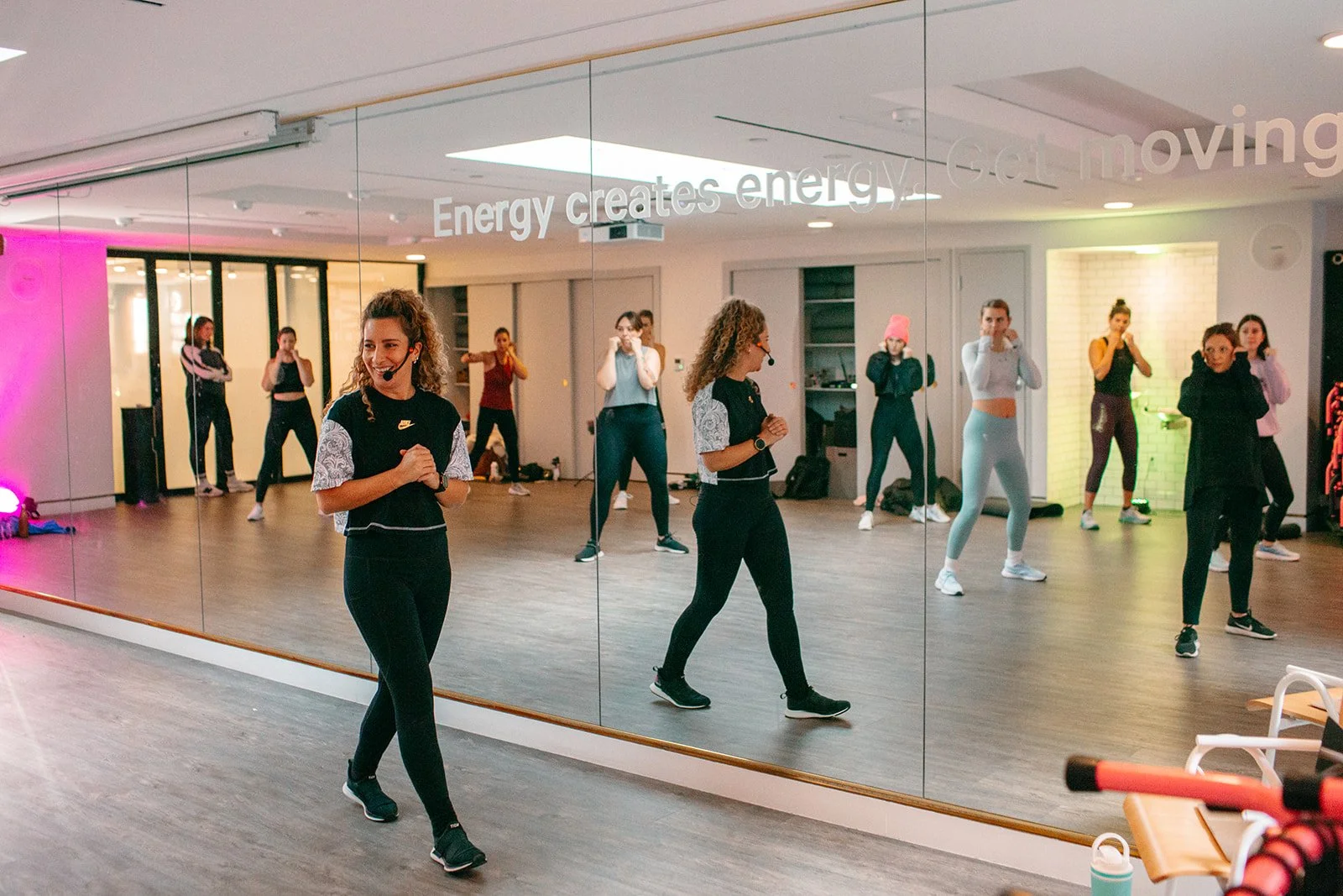 A group fitness class with women exercising in front of a mirror in a gym studio. The instructor is smiling and leading the class, which involves aerobic dance or cardio exercises.