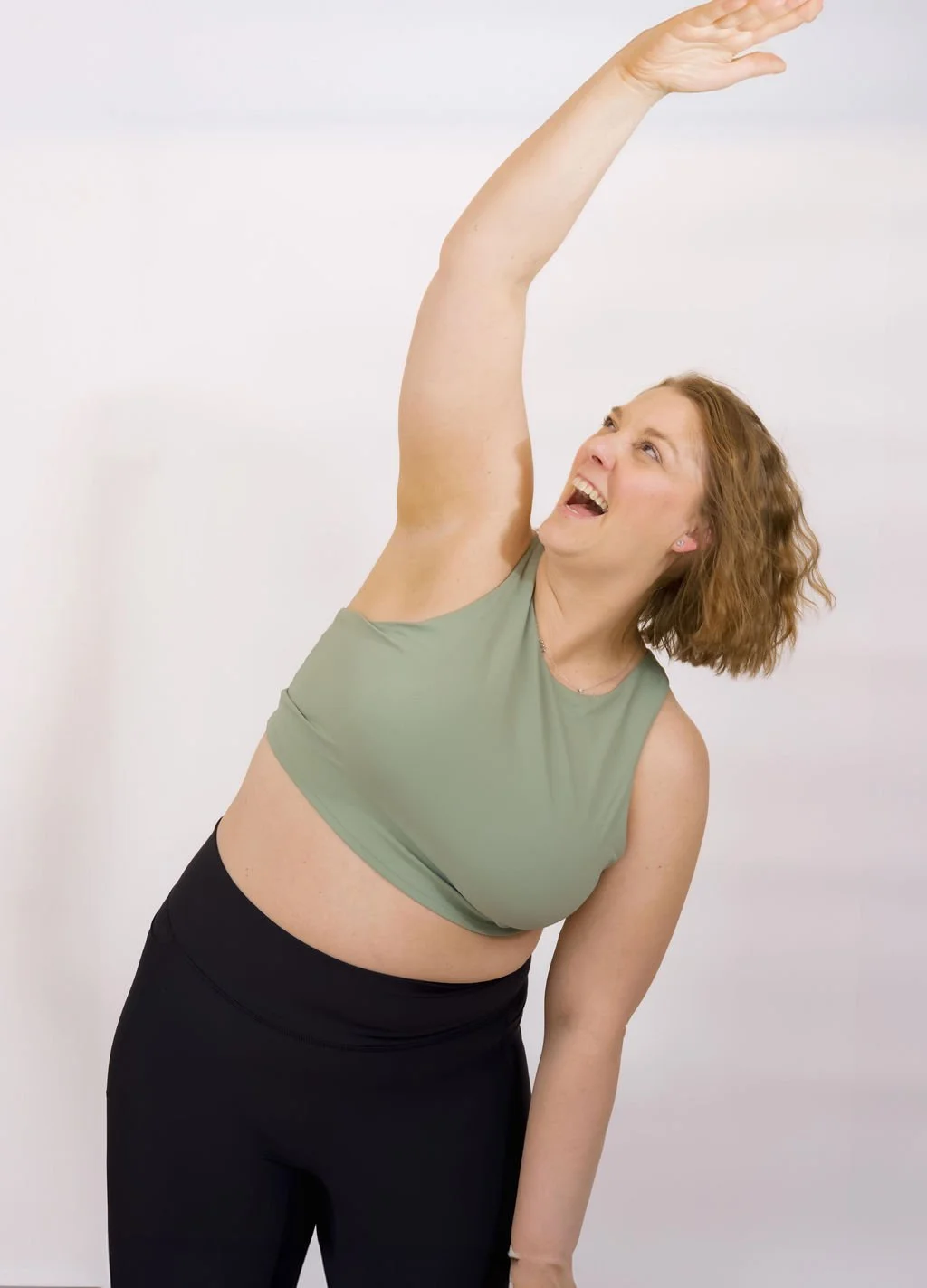 A woman with curly red hair smiling while stretching her right arm overhead, wearing a green sleeveless top and black pants, against a plain white background.