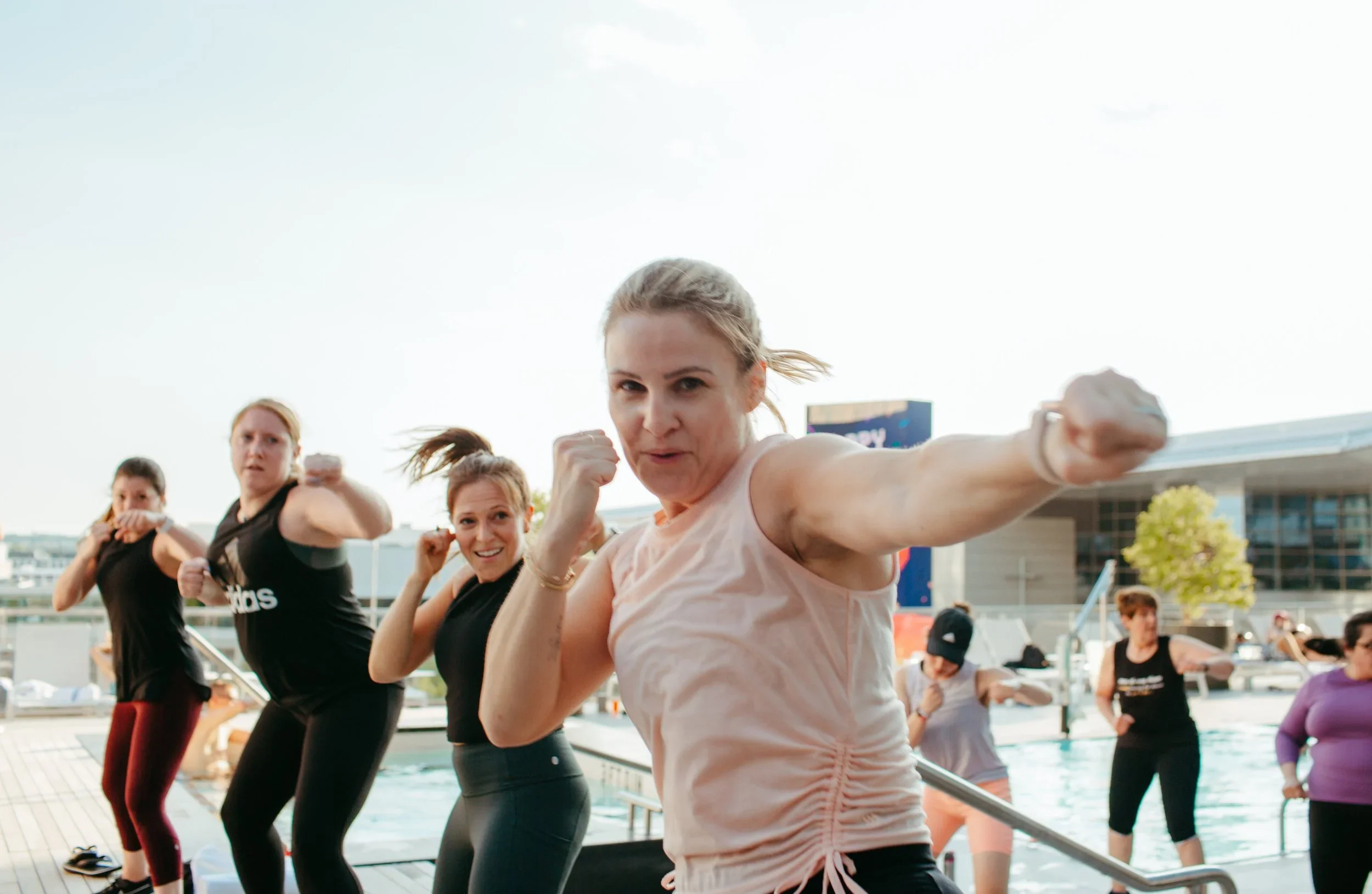 Group of women participating in an outdoor fitness class, performing punches in a synchronized workout, on a poolside area during daytime.
