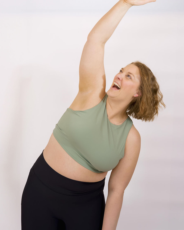 A woman with curly red hair in a green sports bra and black leggings stretching and smiling against a plain white background.