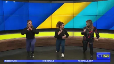 Three women dancing in front of a vibrant, multicolored background on a TV studio set.