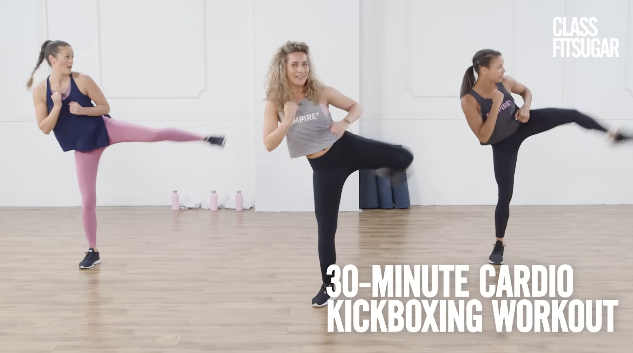 Three women participating in a 30-minute cardio kickboxing workout in a bright, spacious studio with a wooden floor and white walls.
