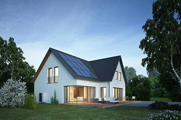 Modern white house with large glass doors, balcony, and solar panels on roof, surrounded by green trees and lawn.