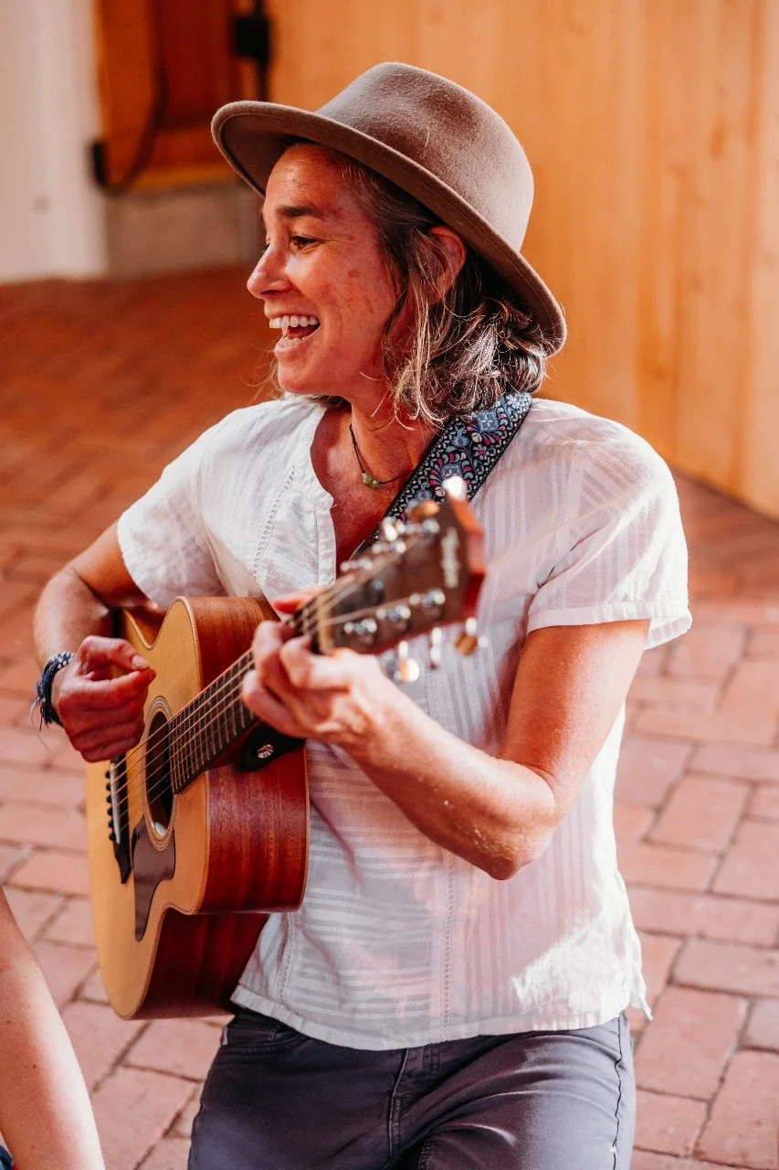 A woman with short hair, wearing a hat, playing an acoustic guitar and smiling indoors with wooden walls and brick flooring.