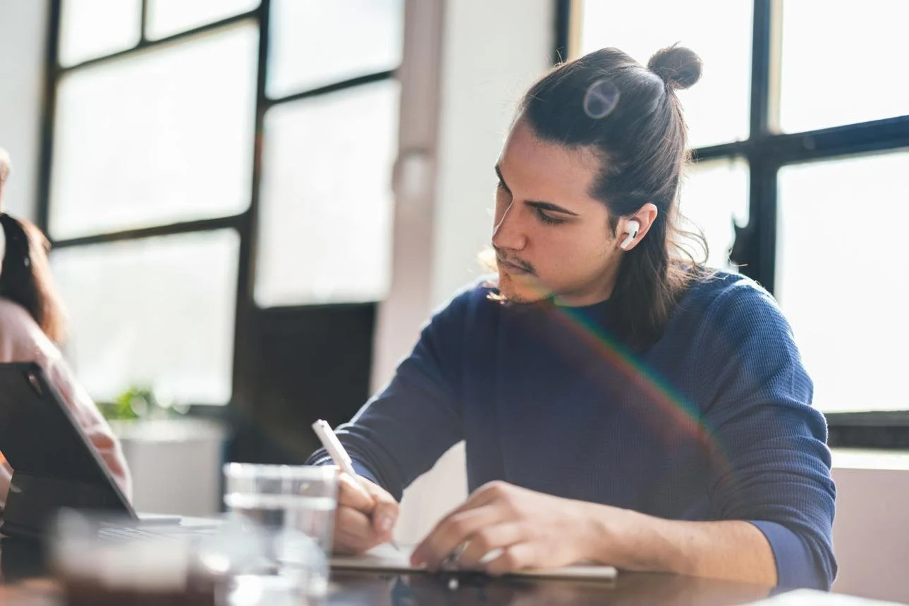 A young man with long dark hair tied in a bun, wearing a blue long-sleeve shirt and wireless earbuds, sitting at a desk, writing in a notebook with sunlight coming through large windows behind him.