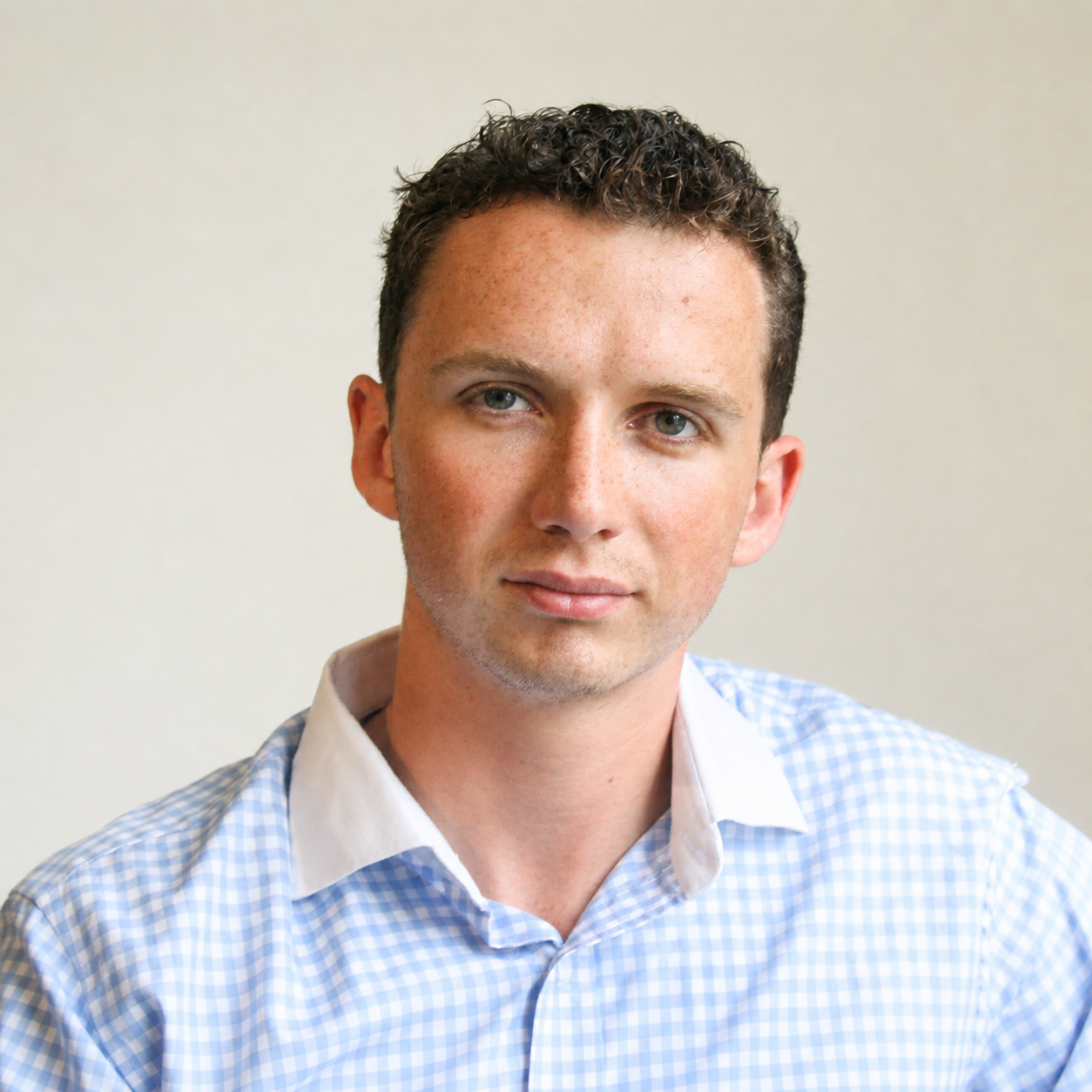 Headshot of a young man with short dark curly hair, blue eyes, wearing a light blue checkered shirt, against a plain beige background.