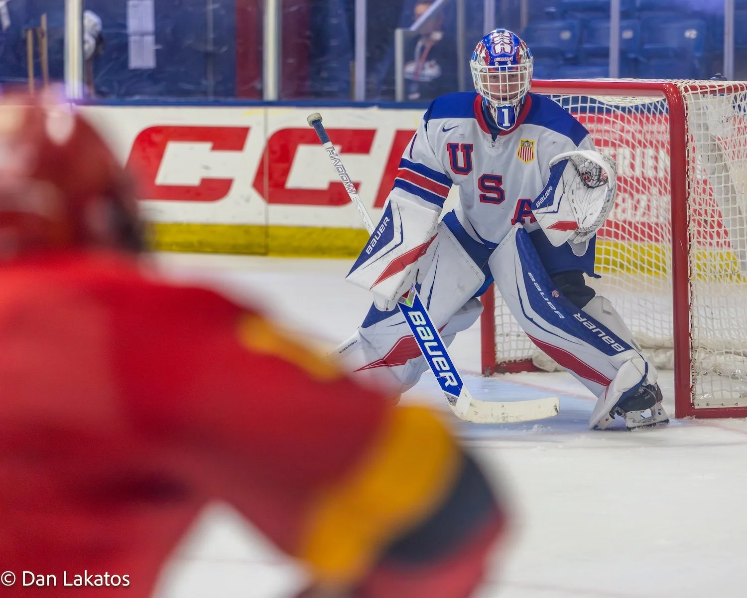 An ice hockey goalie in a USA team uniform making a save on the ice rink, with a blurred player in red in the foreground.
