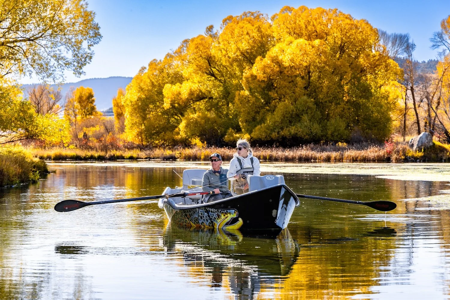 Two people fishing on a boat with a colorful fish design, on a calm river surrounded by autumn trees with yellow foliage. Livingston, Montana, USA