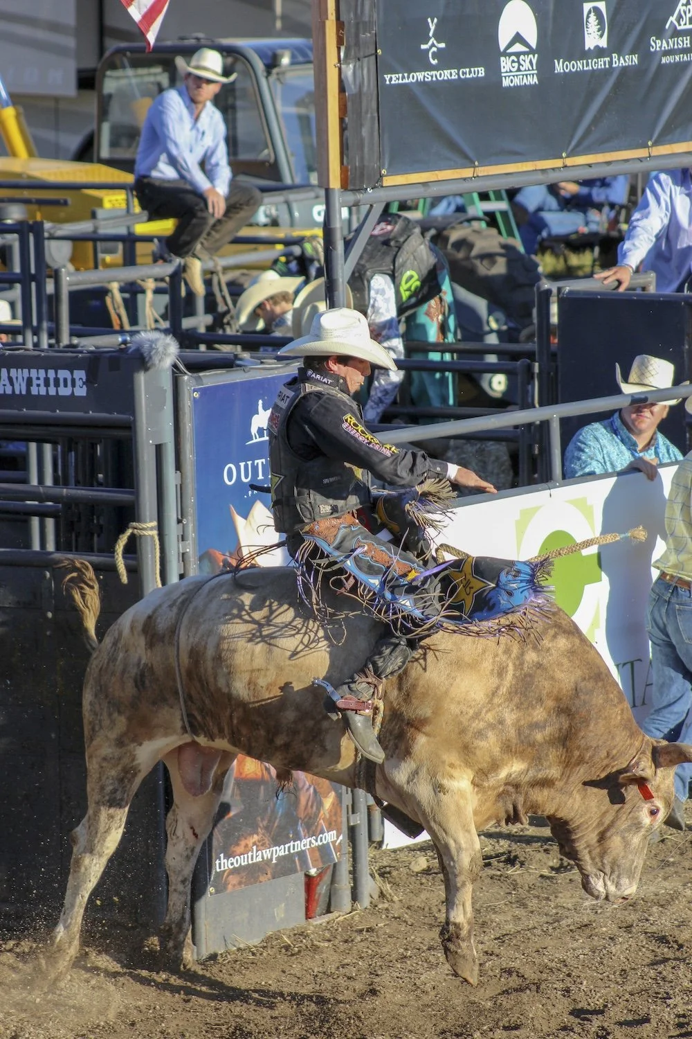 A cowboy riding a bucking bull during a rodeo event, with spectators and officials watching from the sidelines. Pro Bull Rider, PBR, Big Sky, Montana, USA