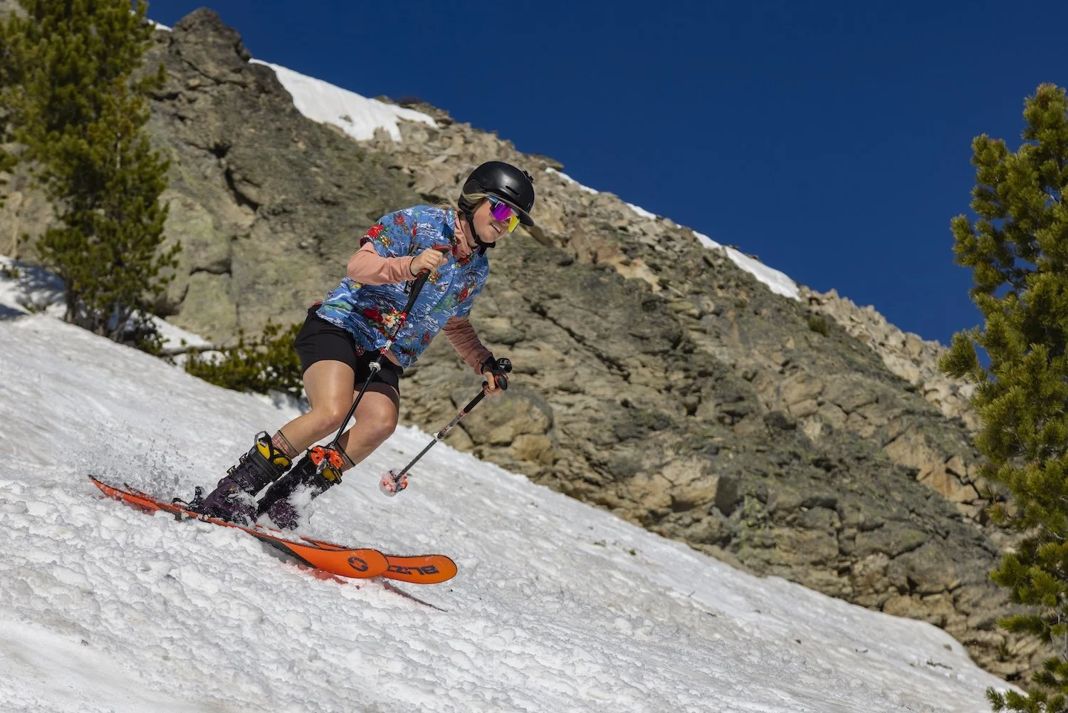 Woman skiing downhill on a snowy mountain slope with rocks and trees around her under a clear blue sky.
