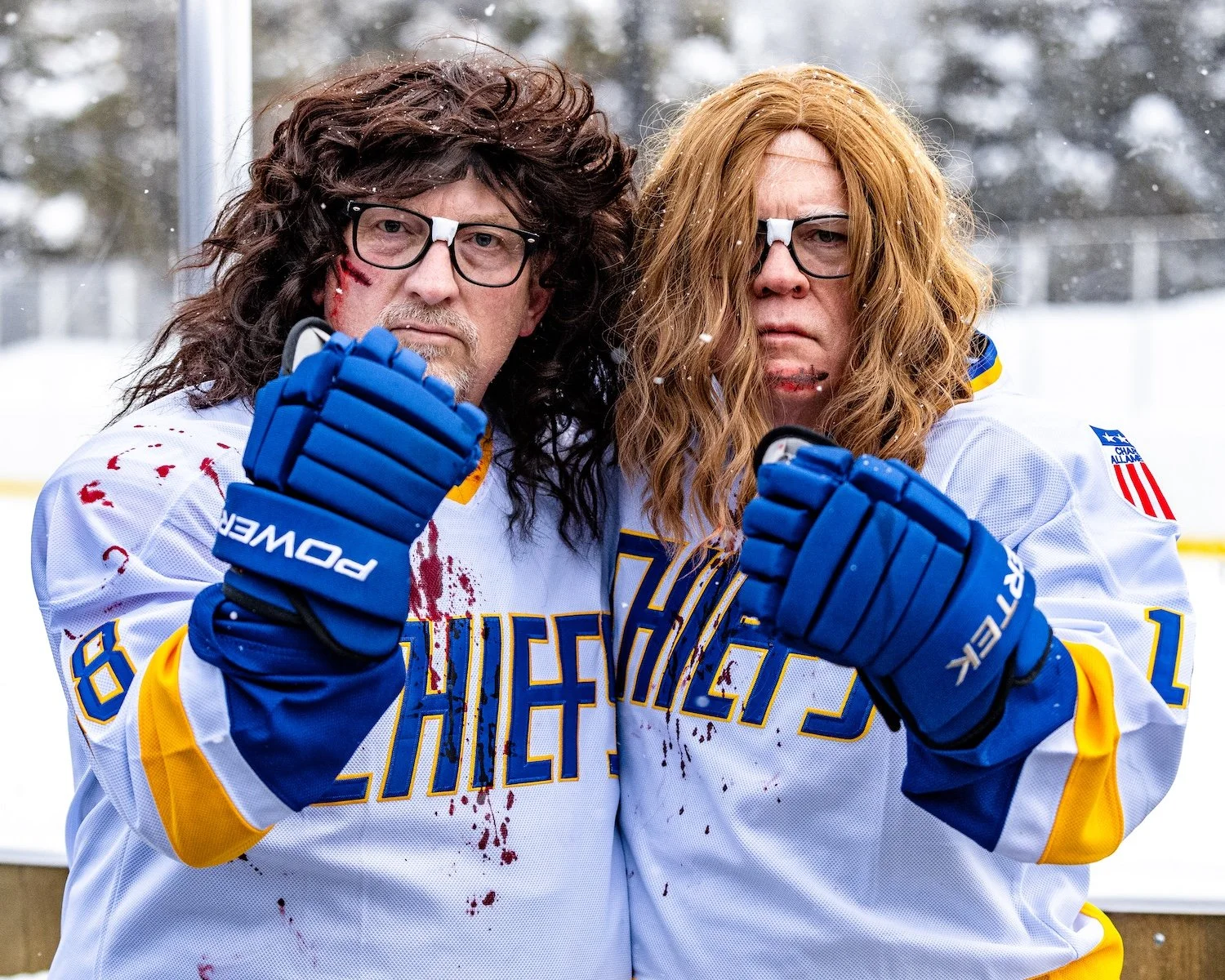 Two hockey players in white jerseys with bloodstains, wearing blue gloves, holding hockey sticks, standing close together outdoors on a snowy ice rink, with expressions of determination.