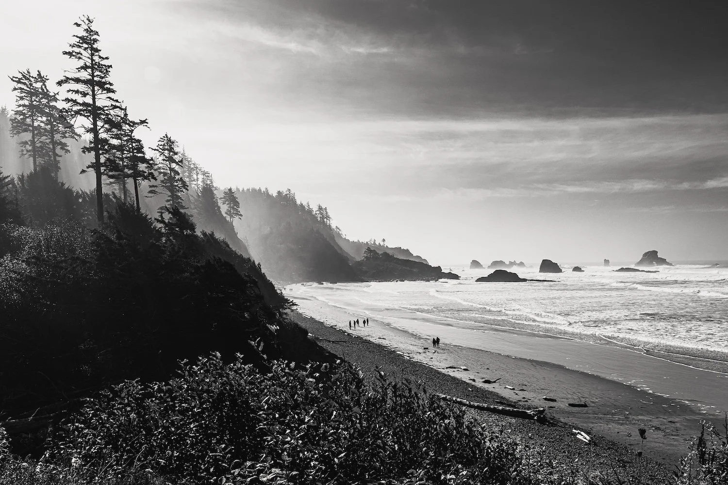 A black and white photo of a beach with cliffs and tall trees on the left, waves crashing on the shore, and several people walking along the beach with rocks in the water in the distance.