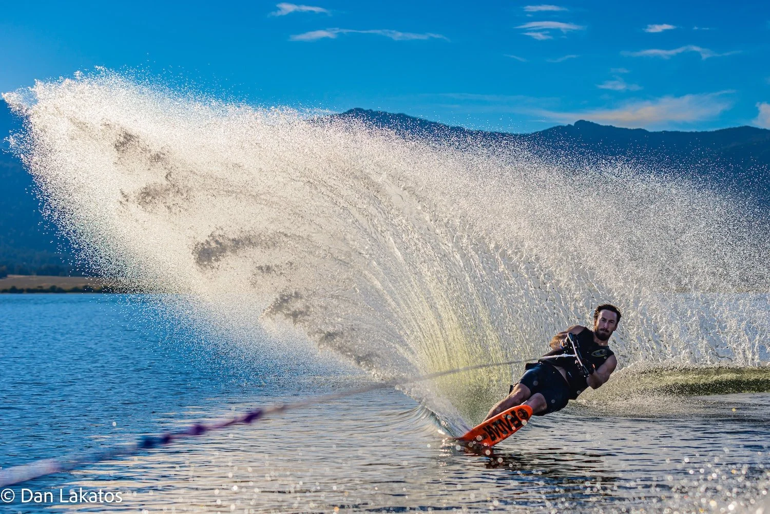 A man water skiing on a lake, creating a spray of water behind him, with mountains in the background and a partly cloudy sky. Hebgen Lake, Montana, USA