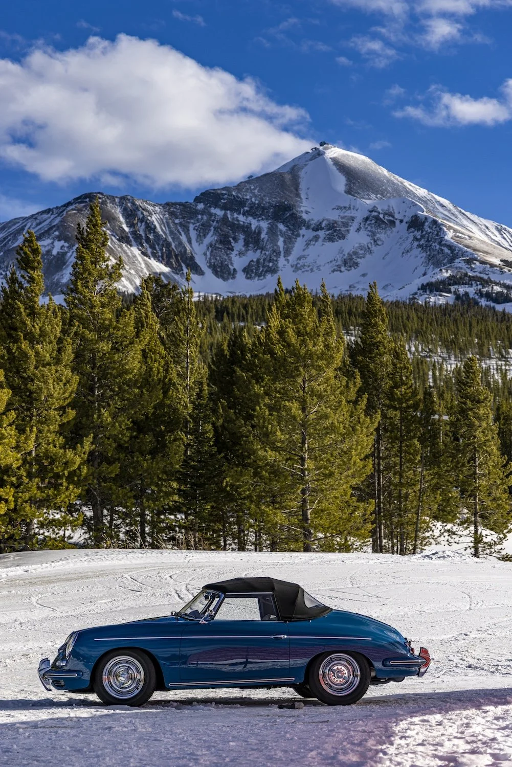 A vintage blue convertible car parked on snow with a backdrop of green pine trees and a snow-capped mountain under a partly cloudy sky. F.A.T. ICE RACE 2026, Big Sky, Montana USA. Dan Lakatos Photo