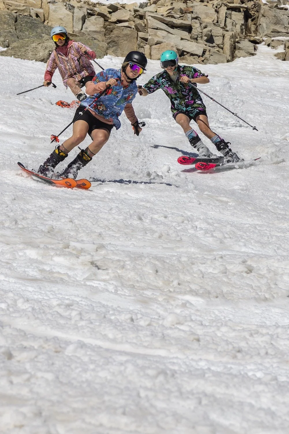 Three women in colorful outfits skiing on a snowy slope with rocks in the background.