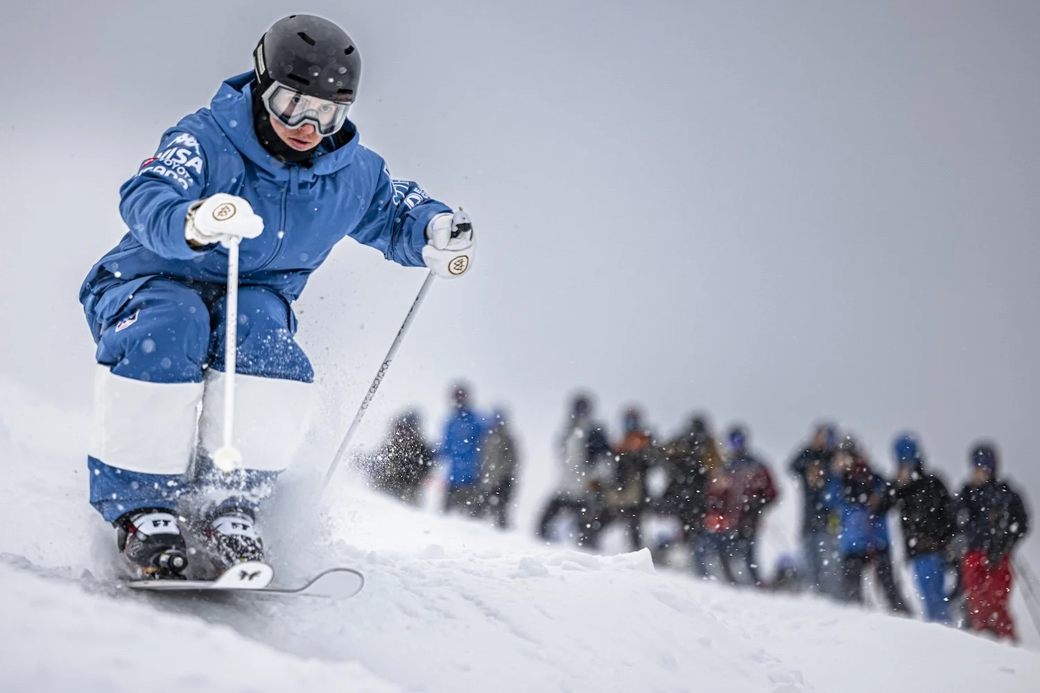 A skier dressed in blue gear navigating a snowy slope during a race, with spectators blurred in the background.