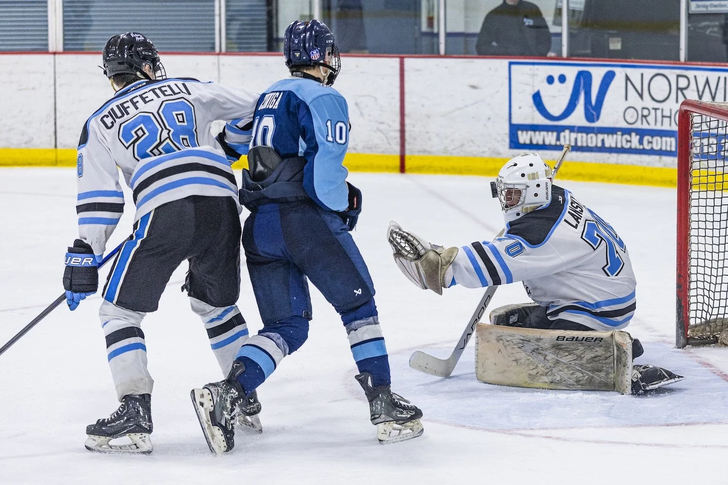 Ice hockey game with two players in white and blue jerseys confronting a player in a blue jersey near the goal, with the goalie in a white jersey attempting to block the puck.