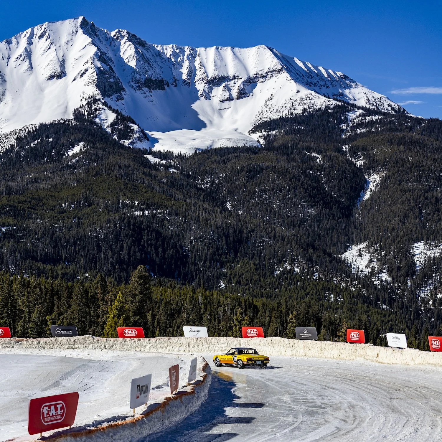 Snow-covered mountain with forested slopes and a small yellow race car on a snowy track in the foreground, with advertising banners along the edge of the track. F.A.T. ICE RACE 2026, Big Sky, Montana USA. Dan Lakatos Photo